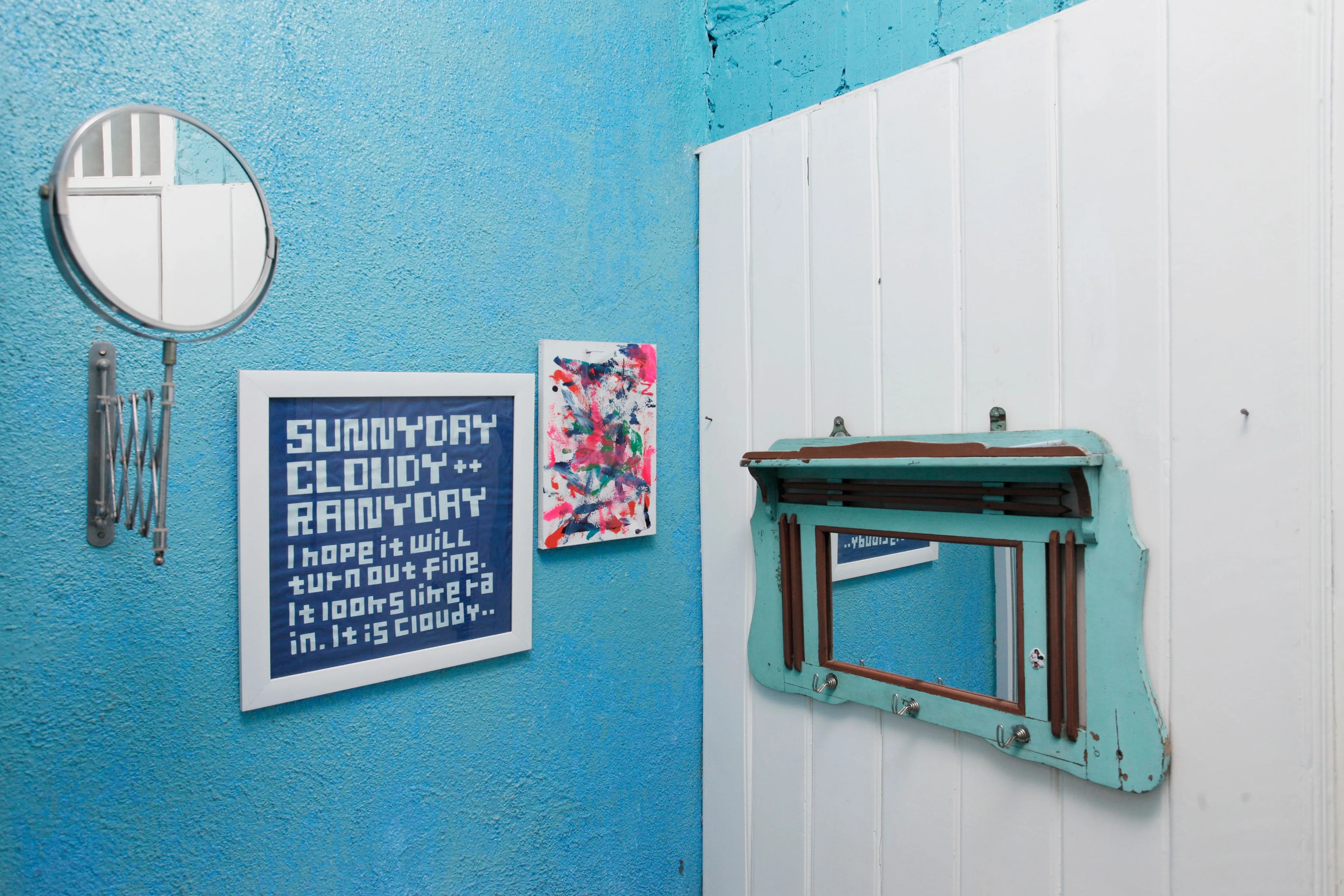 An eclectic turquoise wall with a framed weather-themed text print, a small abstract print, a round mirror and a weathered window frame