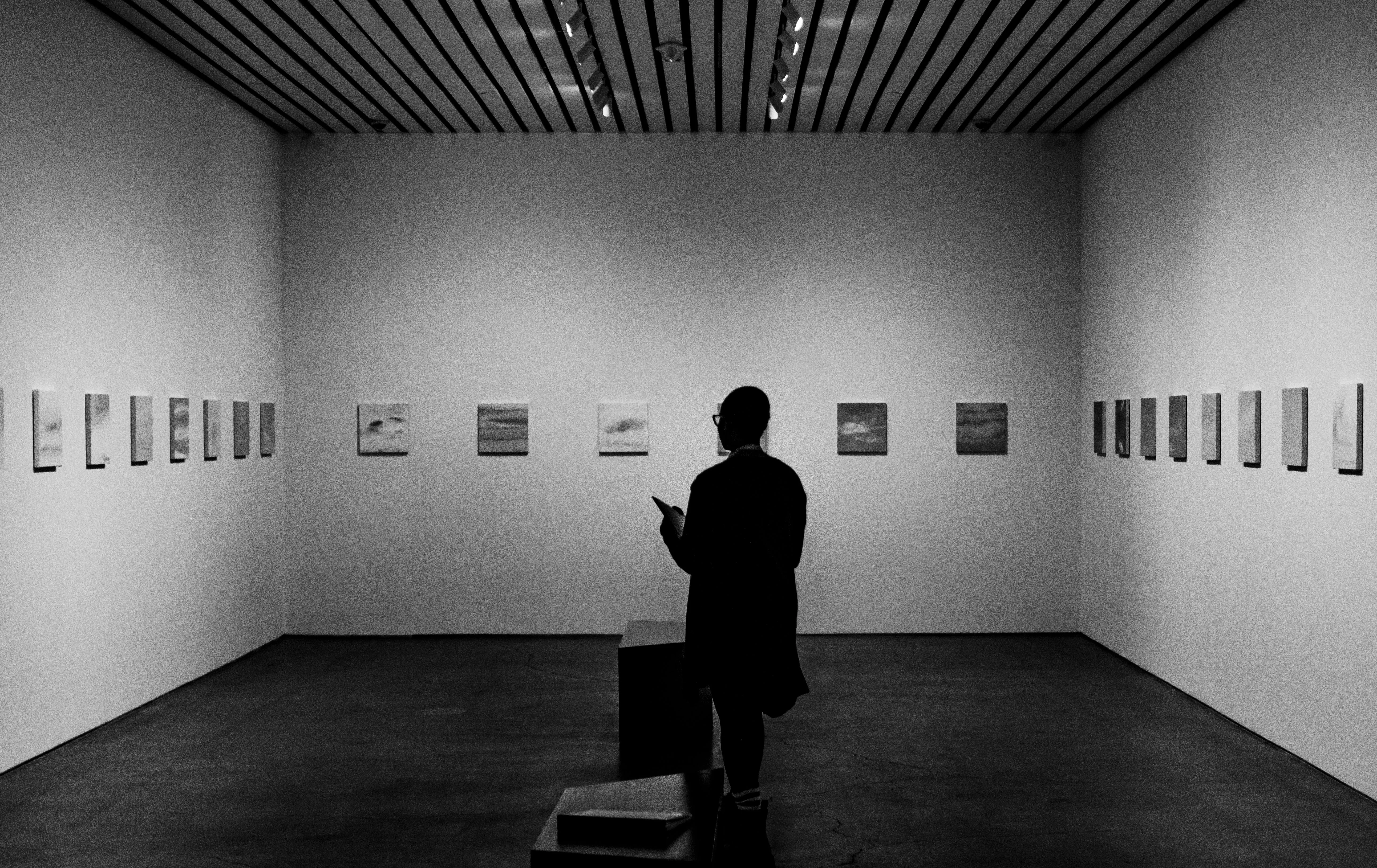 A visitor standing in a quiet minimalist gallery viewing a row of small framed sky-and-sea paintings, black and white