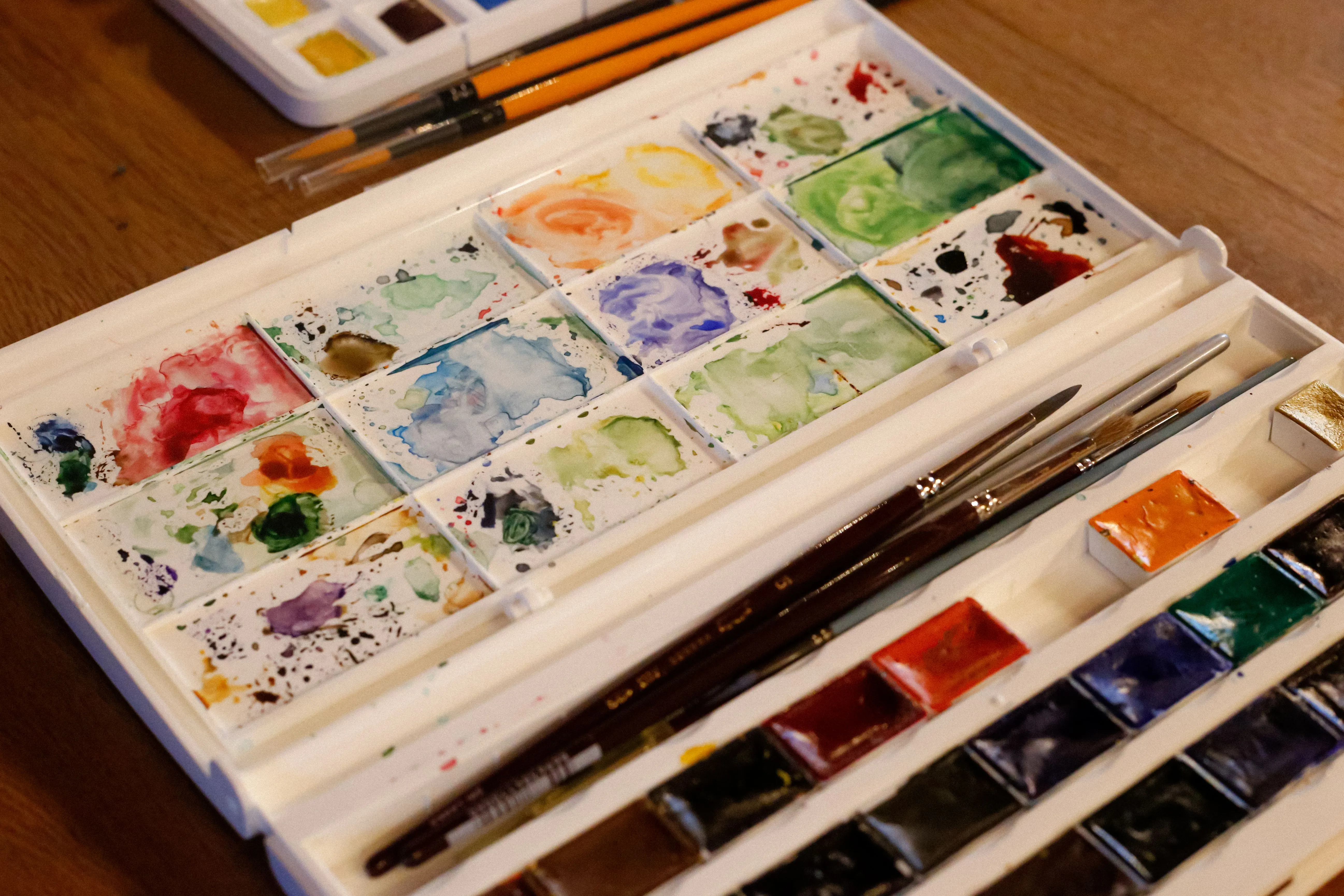 A well-used watercolor palette with stained mixing wells and brushes resting on a wooden table